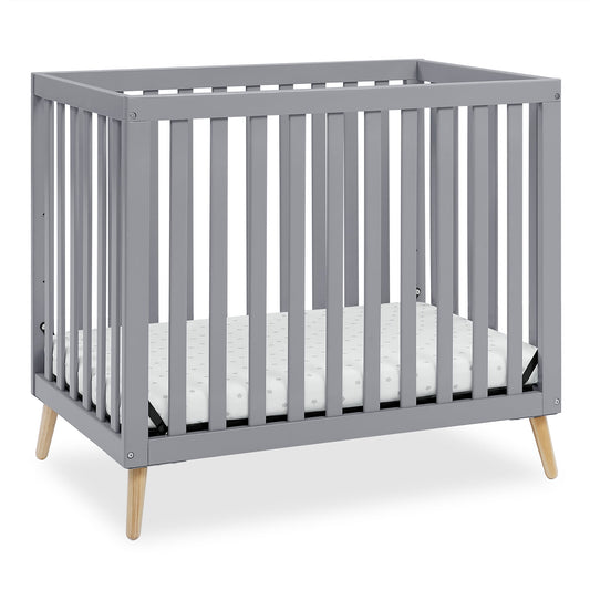 A grey baby crib with a mattress, featuring wooden legs and slatted sides