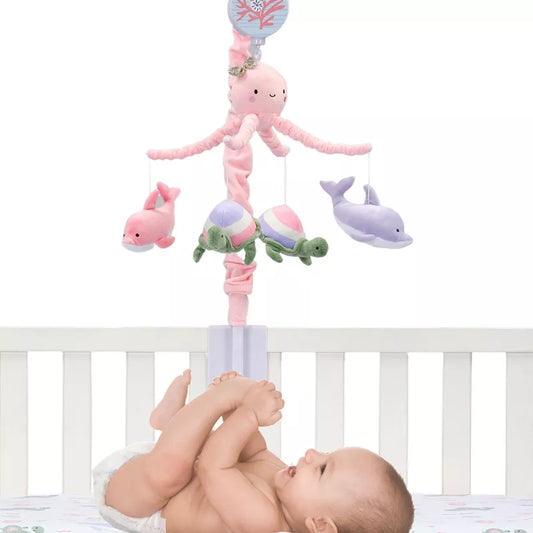 A baby lies in a crib, looking up at a pink musical mobile featuring an octopus, dolphins, and turtles