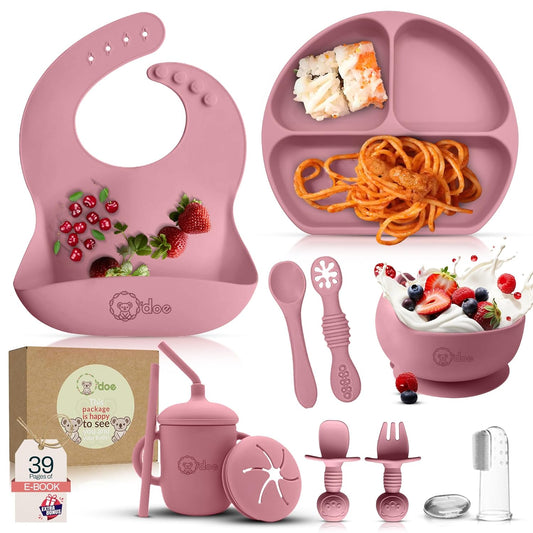 Pink baby feeding set including bib, plate, bowl, cups, and utensils on a white background.