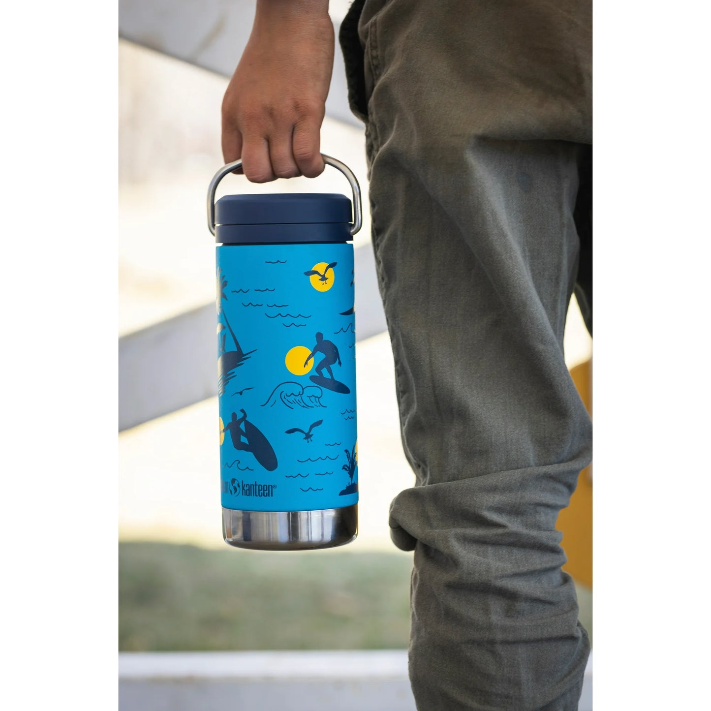 Person holding a blue thermos with illustrations, wearing green pants.