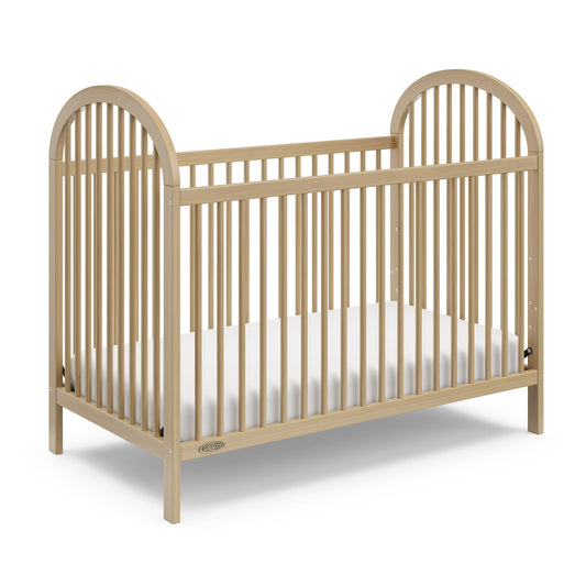 A light-colored wooden baby crib with a white mattress inside