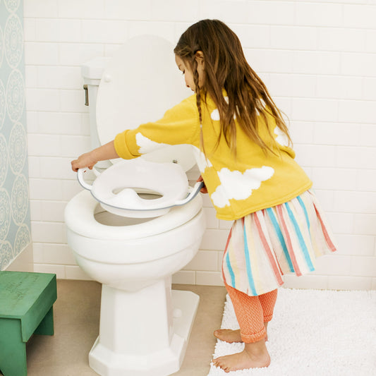 A young child places a white potty training seat onto a toilet in a bathroom setting