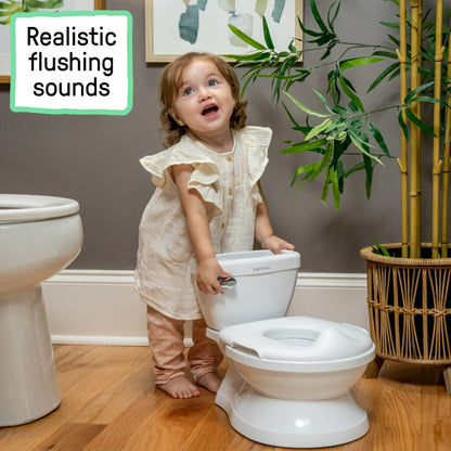 A toddler girl stands next to a white miniature toilet, looking upwards