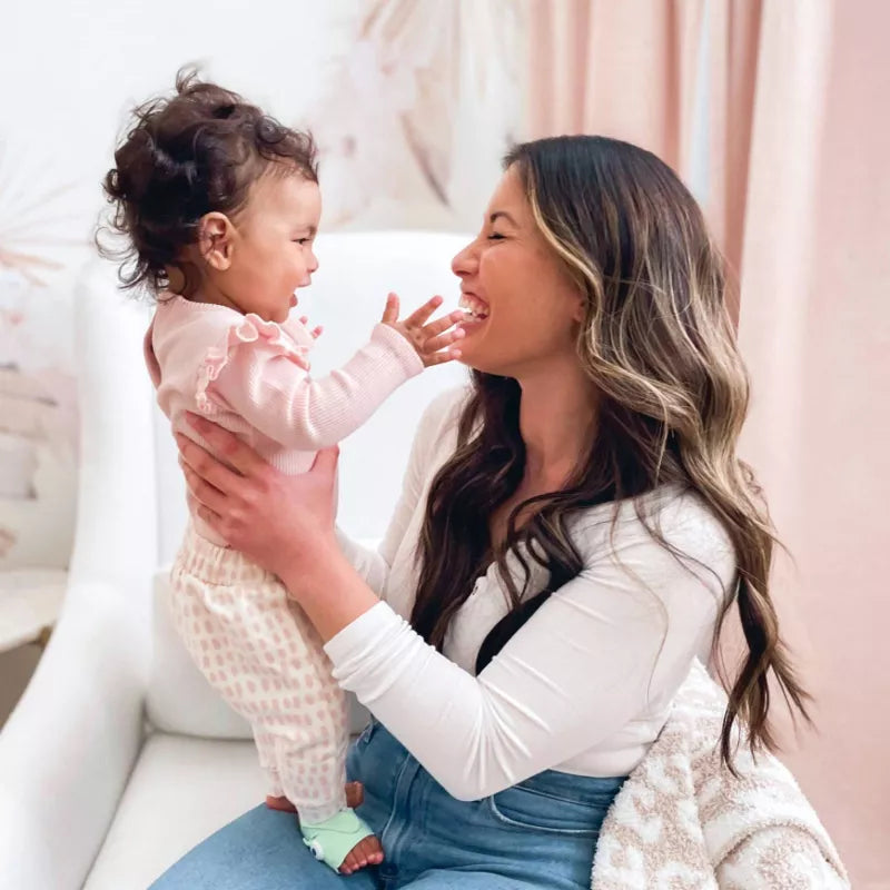 A woman holds a smiling baby, in a light-colored room with curtains