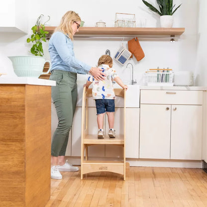 A woman assisting a child on a wooden step stool at a kitchen counter, in a bright, modern kitchen