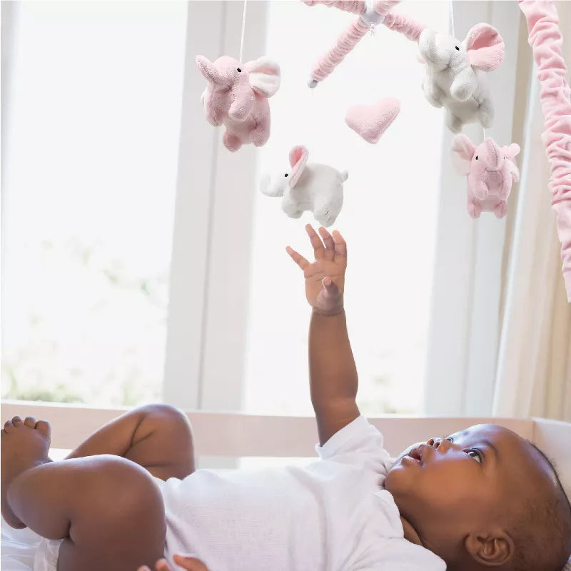 Baby in a crib reaching for a musical baby crib mobile with pink and white elephant toys