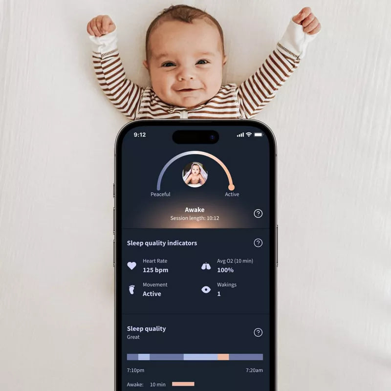 A smiling baby on a bed with a smartphone displaying a baby sleep tracking application
