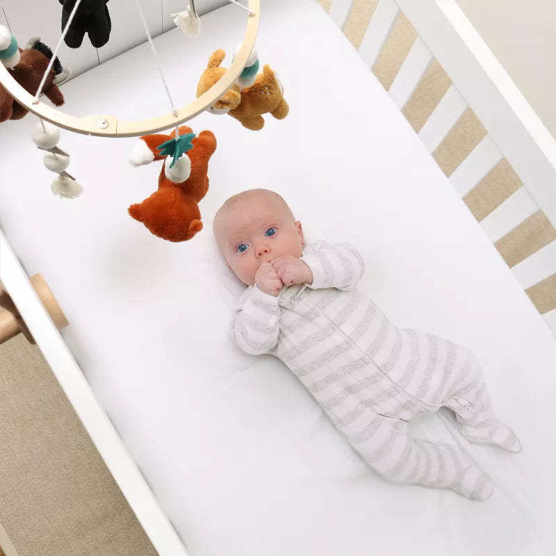 A baby lies in a white crib with a wooden mobile hanging above, featuring animal figures