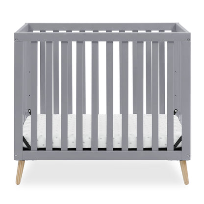 A grey baby crib with a white mattress, featuring natural wood splayed legs