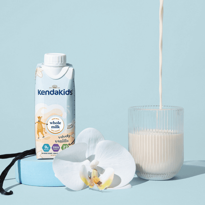 Nutrition shake carton on side with shake pouring into a glass, vanilla bean, and white orchid with blue background