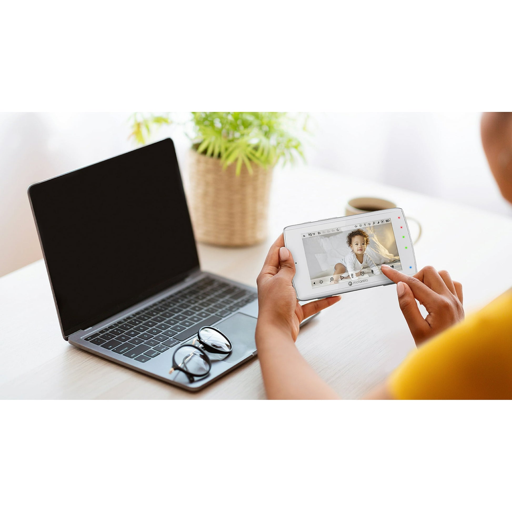A person holds a baby monitor displaying a baby, next to a laptop on a desk